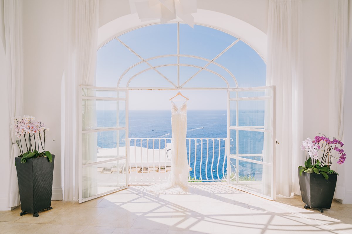 Wedding Dress with a Mediterranean Sea View A stunning wedding dress hangs in an arched doorway with an open sea view, ready for a beautiful villa magia wedding positano