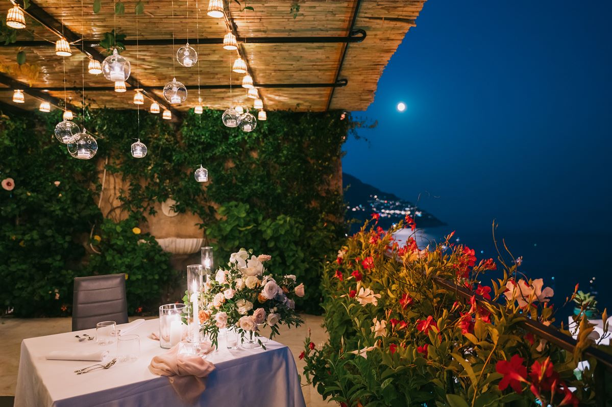 A romantic nighttime table setting for a villa magia wedding positano, with hanging lights, candles, and a full moon over the sea A romantic nighttime table setting for a villa magia wedding positano, with hanging lights, candles, and a full moon over the sea