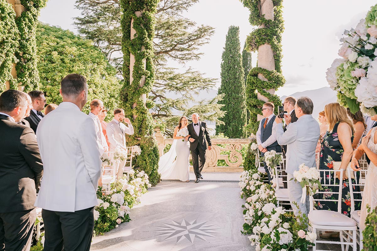 Bride walking down the aisle with her father at a stunning outdoor civil wedding ceremony in Italy, surrounded by elegant floral arrangements