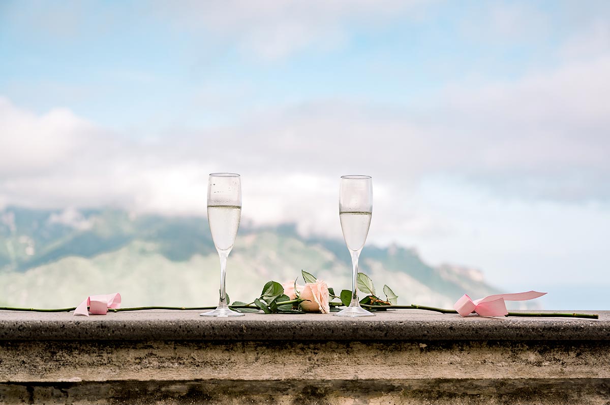 Champagne Toast with a Mountain View Two glasses of champagne and pink roses on a terrace, overlooking the stunning mountain view of a Palazzo Avino wedding Ravello