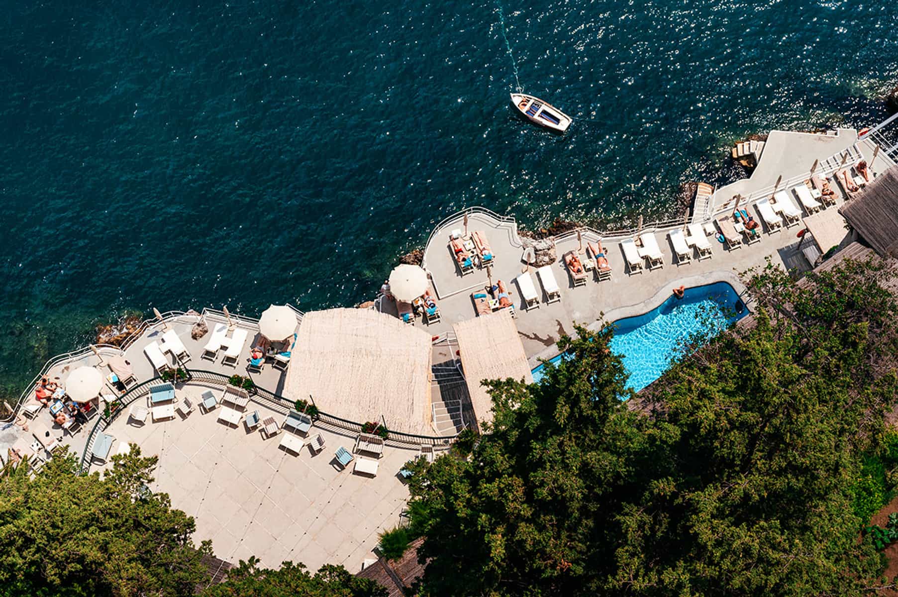A stunning aerial view of a seaside resort on the Italian coast, featuring a pool, sun loungers, and a boat on the deep blue waters: perfect for a wedding experience in Italy
