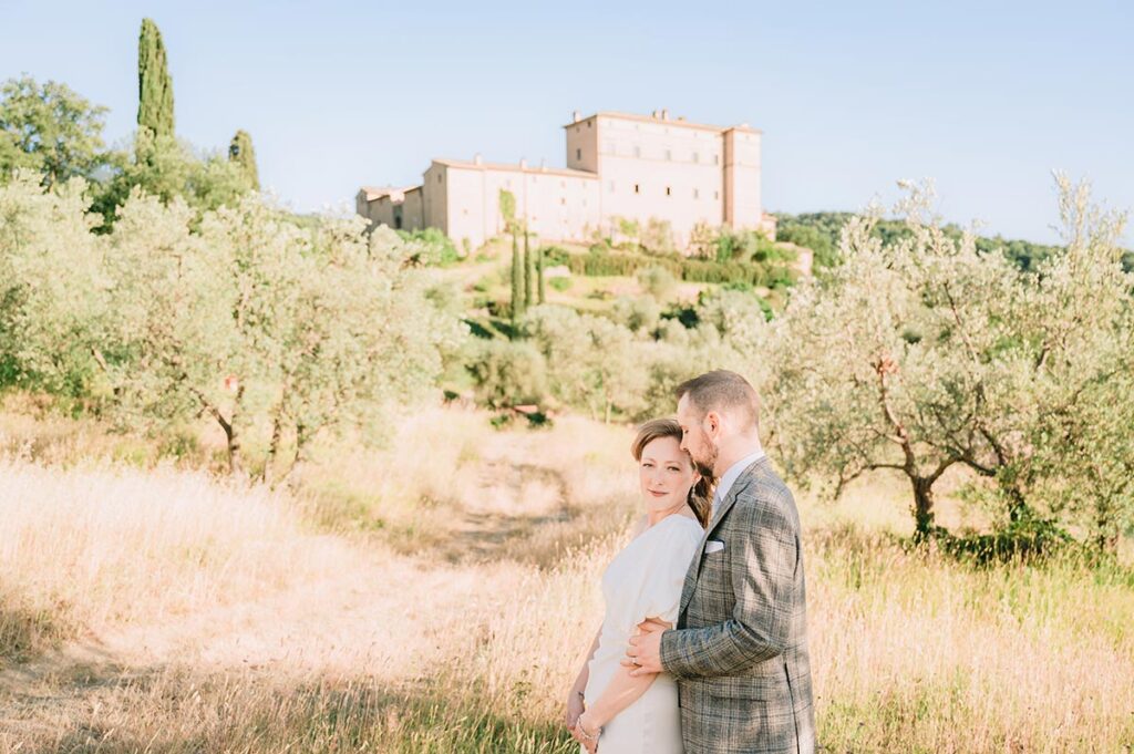 A newlywed couple poses in an olive grove at a Chianti wedding venue, with a historic castle in the background
