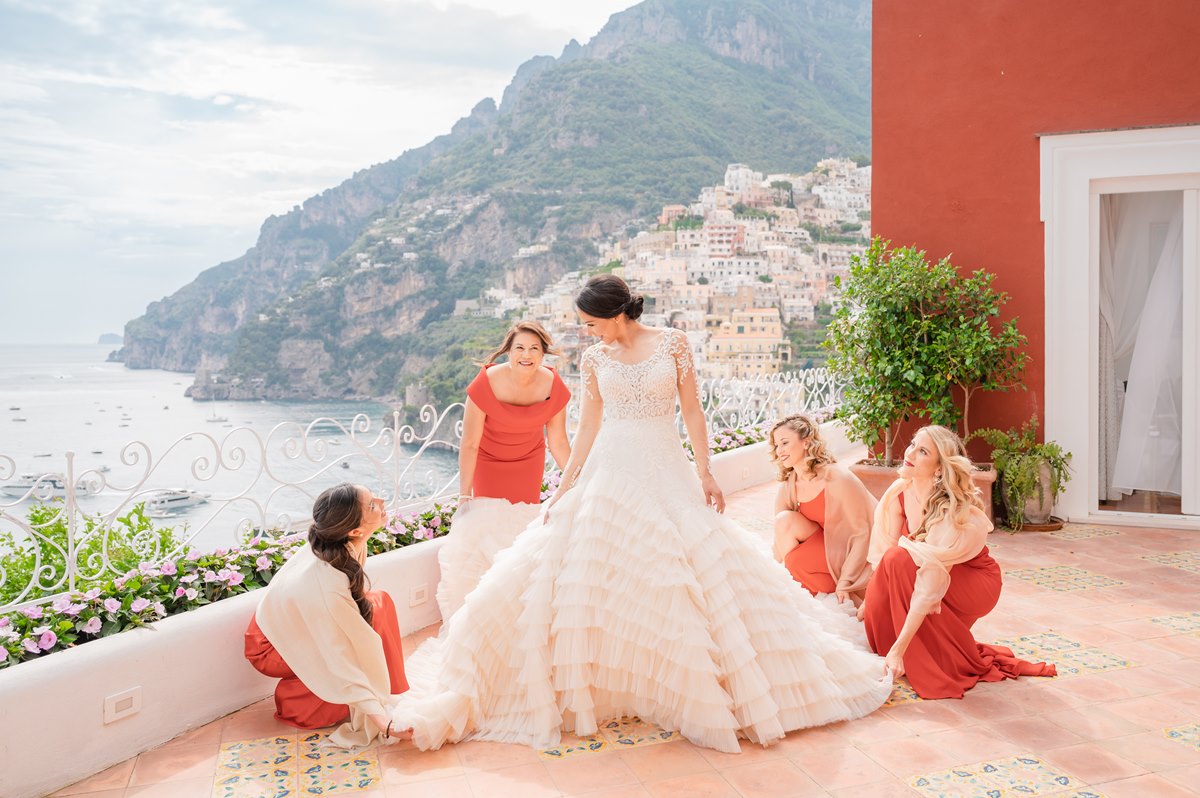 A bride in a stunning gown stands on a terrace overlooking Positano, surrounded by bridesmaids in elegant dresses, capturing a dream wedding experience in Italy.